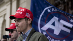 Nick Fuentes wearing a MAGA hat, standing in front of an America First flag.