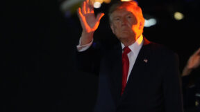 trump waves in the nighttime with a red light shining on him